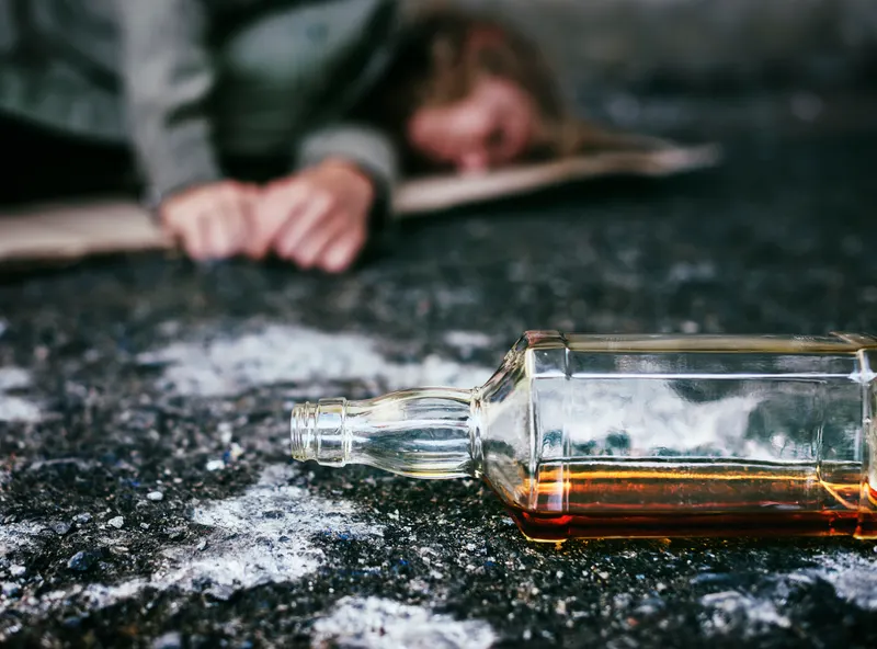 A person slumped on the ground beside an overturned liquor bottle