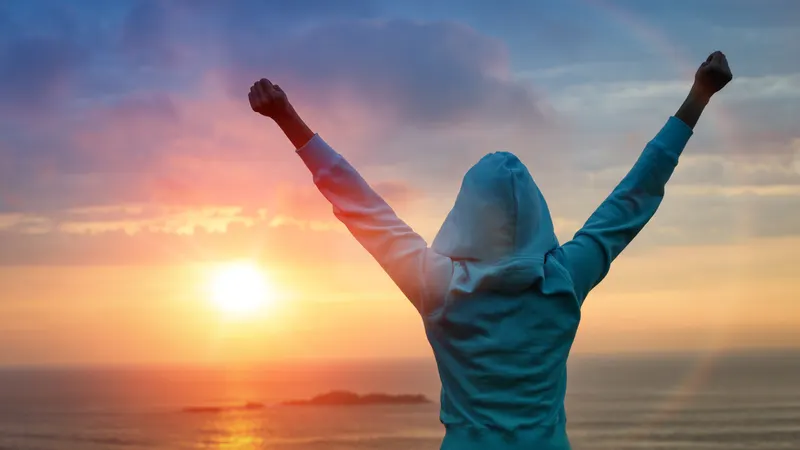 A person in a hoodie raising their arms toward the sunrise over the sea