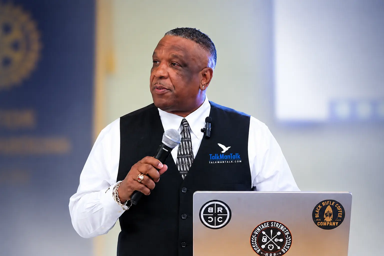 Timothy Stewart speaking into a microphone at a podium in a black TalkManTalk vest and tie